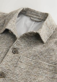 Sacou din lână cu model tartan în nuanțe de bej și maro deschis, cu guler ascuțit, închidere cu nasturi și buzunar pe piept, întins pe o suprafață deschisă.
