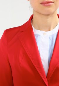 Veste rouge avec des revers pointus, un tissu texturé et une coupe ajustée, portée sur une chemise bleu clair à col avec un détail bouton.