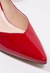 Escarpin rouge en cuir verni avec un bout pointu et un design en V. Texture lisse avec une finition brillante et un intérieur beige.