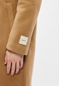 Χέρι resting στο πλάι φορώντας ενός καμηλό χρώματος μαλλινό παλτό με μία λευκή ετικέτα Calvin Klein στον μανίκι.