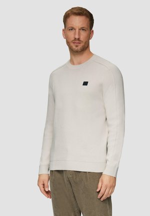 Lichtbeige trui met een ronde halslijn en geribbelde manchetten, voorzien van een klein donker logo-opdruk op de borst en een gladde gebreide textuur.