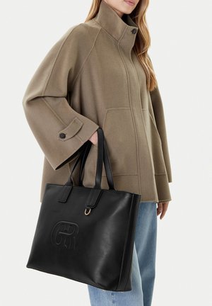 Mujer con abrigo marrón claro de cuello alto y jeans azules, sosteniendo un gran bolso tote negro de cuero con logo en relieve y asas dobles.