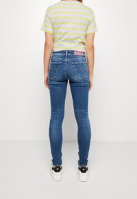 Dopasowane niebieskie dżinsy z tylnymi kieszeniami, delikatne przetarcia i kontrastujące przeszycia; w połączeniu z szaro-limonkową koszulką w paski i białymi butami.