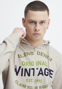 Krémová zipsová mikina vyrobená z mäkkej tkaniny, so silnými textovými vzormi v zelenej a námorníckej farbe s nápisom "VINTAGE" a "CLAIM YOUR RIGHT."