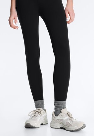 Ben iført sorte leggings, grå ribstrikkede sokker og off-white chunky sneakers, stående på en lys overflade.