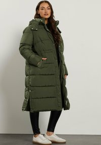 Lang, olivengrøn pufferjakke med hætte, der har trykknapper, sidelommer og en quiltet struktur. Sammen med sorte leggings og hvide sko.