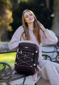 Černý batoh s růžovými detaily, vybavený designem s pružným kabelem, kapsami na zip a nastavitelnými popruhy, položený na dřevěné lavičce.