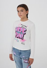 T-shirt bianco a maniche lunghe con una grafica colorata che recita "ECCO CHE ARRIVA IL DIVERTIMENTO!" con accenti rosa e blu. Indossato con jeans blu strappati.