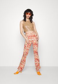 Top curto bege com acabamento brilhante, combinado com calças de padrão ondulado multicolorido. A modelo usa óculos de sol laranja e saltos a condizer.