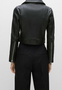 Sort cropped læderjakke med synlig tekstureret finish, stor krave og sølv lynlåse ved ærmeåbningerne. Har et logo ved baghalen.