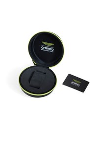 Funda circular negra con cierre verde, que contiene un artículo negro con textura suave y una tarjeta con marca. El forro interior es negro.