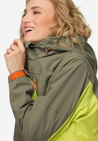 Chaqueta impermeable en color oliva y amarillo brillante, con un alto cuello, detalles ajustables en naranja y un bolsillo frontal con cremallera.