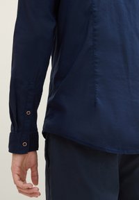 Mörkblå skjorta i lätt tyg med långa ärmar, knappade manschetter och en rundad fåll. Har en slät textur och subtil lyster.