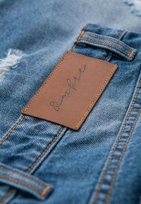 Denim material med en slitagen look; har en brun läderetikett sydd med ordet "Ombre" präglat. Ljusblå färg, strukturerad yta.