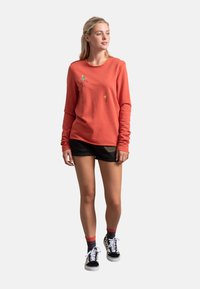 Felpa rossa a maniche lunghe con dettagli ricamati minimalisti, abbinata a pantaloni corti neri e sneakers a fantasia. Tessuto morbido, vestibilità casual.