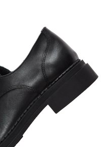Chaussure en cuir noir au fini lisse, à bout rond, avec un petit talon et des détails de coutures visibles le long de l'empeigne et de la semelle.