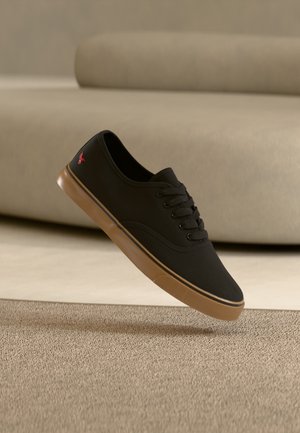 Baskets basses noires avec semelle en caoutchouc marron et lacets, flottant au-dessus d'un tapis beige et d'un canapé beige clair en arrière-plan.