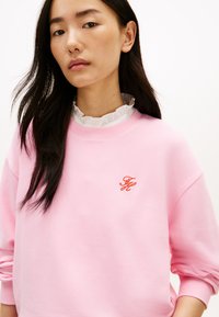 Ροζ φούτερ με στρογγυλή λαιμόκοψη, που διαθέτει λευκή βολάν γιακά από κάτω. Εμφανές κόκκινο κεντημένο λογότυπο στο στήθος.