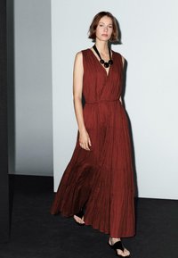 Model walks confidently in a deep red, sleeveless maxi dress, paired with a bold black necklace, against a minimalist backdrop.