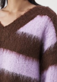 Gros plan d'une personne portant un pull en maille duveteuse marron et violet clair avec un col en V.