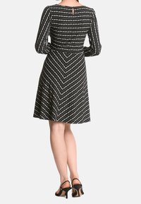 Schwarzes Kurzarmkleid mit weißem Zickzack-Muster, tailliertem Oberteil, ausgestelltem Rock und Schlüsselloch-Detail im Rücken. Akzentuiert mit schwarzen Absatzsandalen.