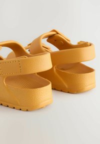 Gelbe Sandalen mit einer strukturierten Gummibeschichtung, ausgestattet mit verstellbaren Riemen und einem erhöhten Absatz. Entwickelt für Komfort und Rutschfestigkeit.