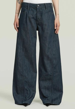 Szerokie ciemnoniebieskie dżinsy z denimu noszone przez osobę stojącą z ramionami swobodnie opuszczonymi wzdłuż boków, z widocznym przednim guzikiem i kieszeniami.