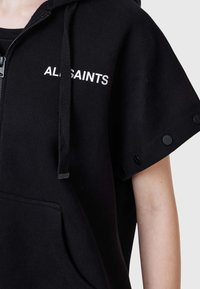 Sudadera con capucha negra de tela suave, con cremallera frontal, mangas oversize con detalles de botones y el logo "ALLSAINTS" en blanco.