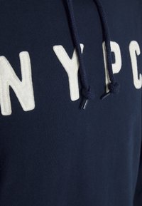 Sudadera azul marino con grandes letras de felpa blanca "N", "Y", "P", "C" en la parte frontal. Cuenta con cordones de textil azul marino con puntas de metal.