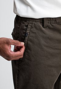 Donkergroene, lichte broek met een elastische tailleband en een zijzak. Heeft een klein logotag op de zak. Zachte, textuurachtige stof.