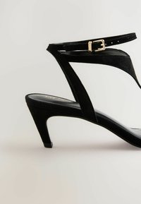 Sandalini con tacco alto in suede nero, caratterizzati da un sottile cinturino alla caviglia, punta appuntita e un tacco leggermente curvo per una vestibilità sicura.