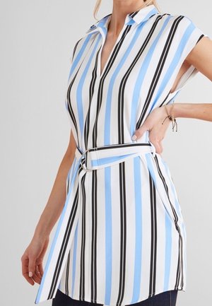 Femme portant une tunique sans manches ceinturée avec des rayures verticales noires, blanches et bleu clair, debout la main sur la hanche.