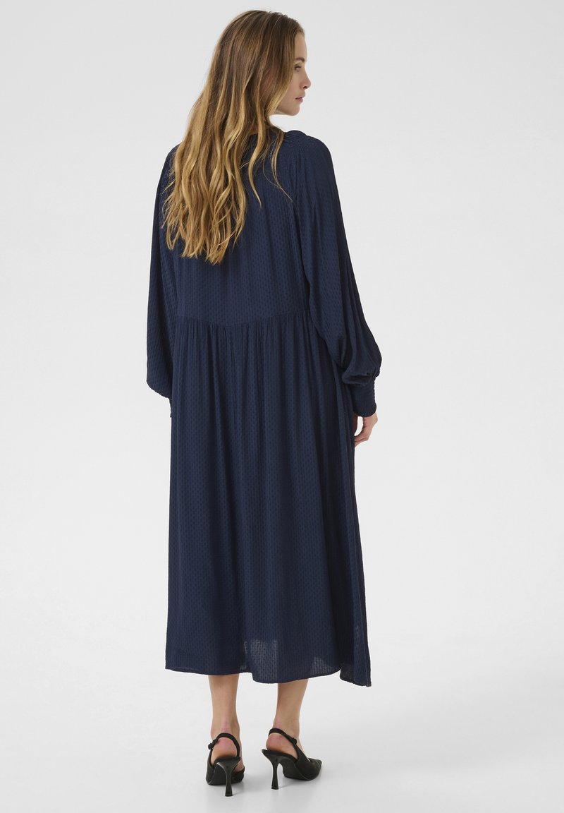 Marineblaues, langärmeliges Midi-Kleid aus strukturiertem Stoff, mit tailliertem Oberteil und schwingendem Rock. Tragen mit schwarzen Absatzsandalen.