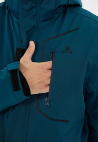 Hand gleitet in die vertikale, mit Reißverschluss versehene Brusttasche einer teal-farbenen, wasserdichten Jacke mit schwarzem Logo und einem Klettverschluss am Handgelenk.