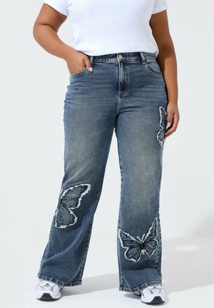 Person, die weite blaue Jeans mit drei großen Schmetterlingsaufnähern an den Beinen trägt, kombiniert mit weißen Turnschuhen und einem weißen Hemd.