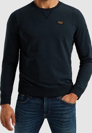 Mann trägt dunkelblaues Sweatshirt mit langen Ärmeln und kleinem Brustlogo und blaue Jeans, Hand in der Tasche, vor einfachem Hintergrund.
