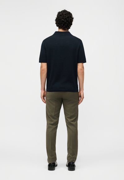 Άνδρας με σγουρά μαλλιά στέκεται με την πλάτη, φοράει μαύρο πόλο με κοντά μανίκια, χακί παντελόνι και μαύρα παπούτσια σε λευκό φόντο.