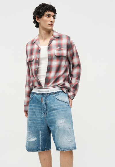 Άνδρας με κόκκινο και γκρι καρό πουκάμισο πάνω από λευκό αμάνικο, συνδυασμένο με ανοιχτό μπλε σκισμένα τζιν σορτς και ορατή ζώνη με λογότυπο.