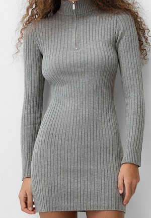 Femme portant une robe en maille grise ajustée, à manches longues et côtelée, avec une courte fermeture éclair au col montant.