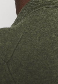 Närbild av en person som bär en texturerad olivgrön tröja med en ribbad krage, som visar området vid bakre axeln.