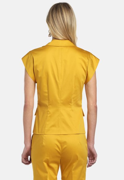 Szőke nő háttal a kamerának, szűk sárga rövid ujjú blézert és hozzá illő sárga nadrágot visel.