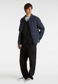 Marineblauwe textuur blazer met knopen, zwarte ritssluiting vest met zakken, zwarte cargobroek en geruite instap schoenen.