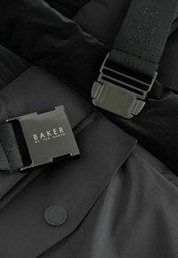 Melnā pūkaina jaka ar taisnstūrveida metāla sprādzi, uz kuras ir iegravēts "BAKER by TED BAKER", un teksturēti auduma akcenti.
