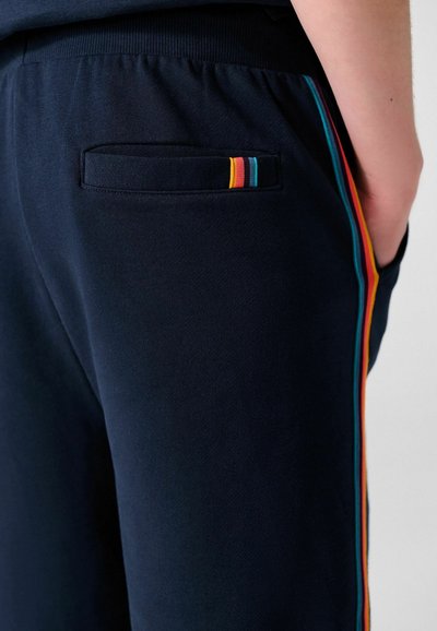 Pantalones azul marino con costuras laterales a rayas de arcoíris y un bolsillo trasero, usados con la mano apoyada en el bolsillo delantero.