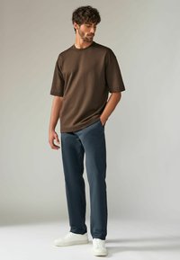 Camiseta de algodón marrón con mangas cortas, combinada con pantalones azul marino y zapatillas blancas. Diseño minimalista, corte relajado, sin patrones visibles.