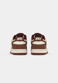 Par noguće Nike smeđe i bež boje prikazane sa stražnje strane, s naglaskom na logo na potplatu protiv jednostavne bijele pozadine.