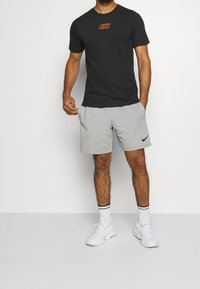 Černé tričko Nike s oranžovým logem, v kombinaci se šedými šortkami s černým swoosh Nike, a bílými teniskami s kontrastními černými akcenty.