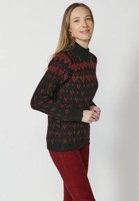 Pull en tricot noir et rouge à motifs avec un col haut, présentant un design texturé et des poignets côtelés. Le mannequin porte un pantalon rouge.