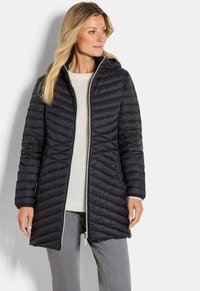 Vrouw in een zwart gevoerd hoodie-jack over een witte trui en grijze broek, kijkend naar rechts tegen een effen achtergrond.