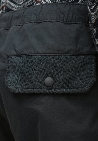 Czarne spodnie z tkaniny z teksturowaną kieszenią z patką o jodełkowym wzorze, zamykaną na okrągły czarny guzik. Ogólnie gładka faktura.